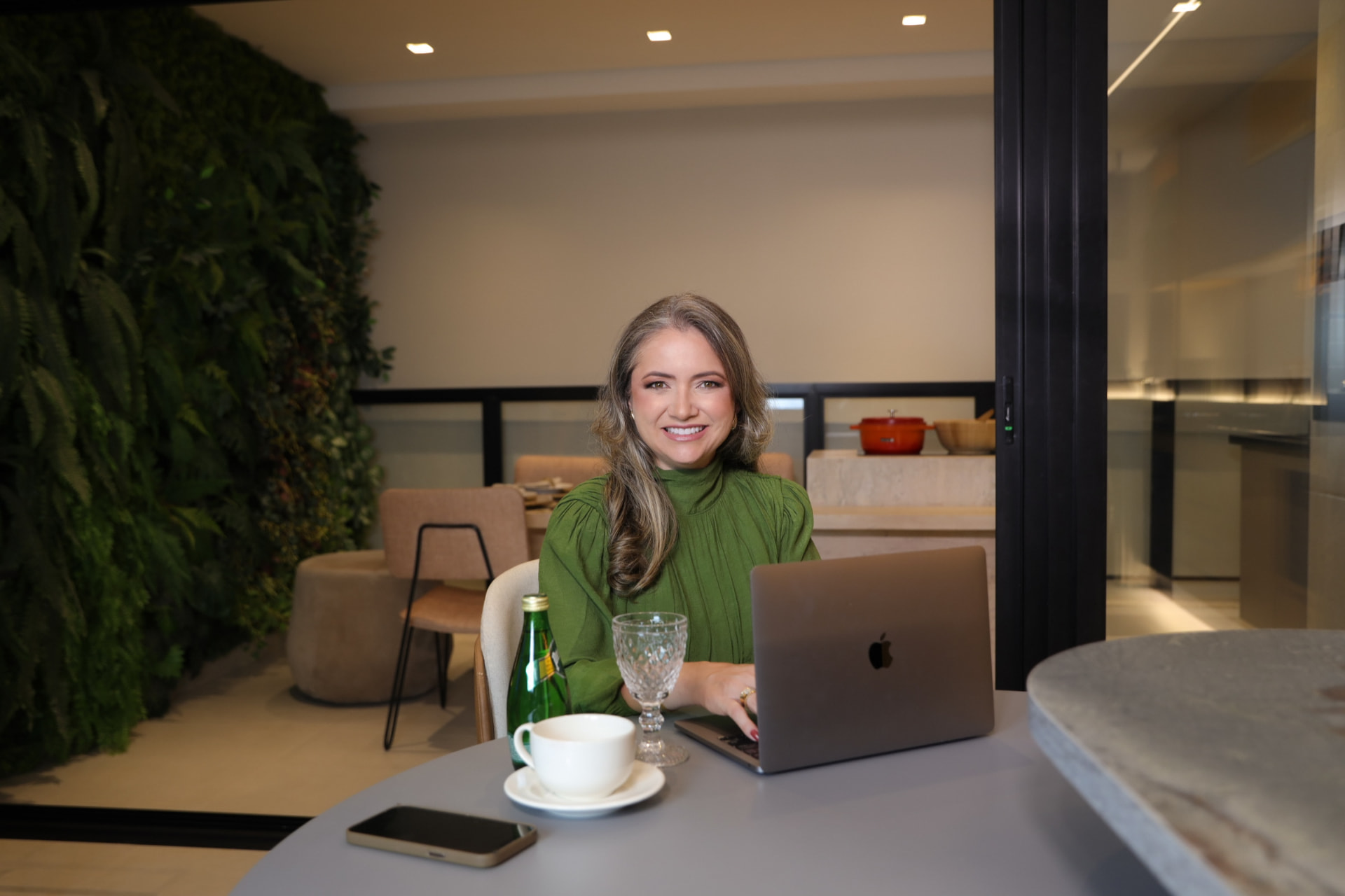 Rejane Duarte sentada à mesa, sorrindo, em ambiente com parede verde