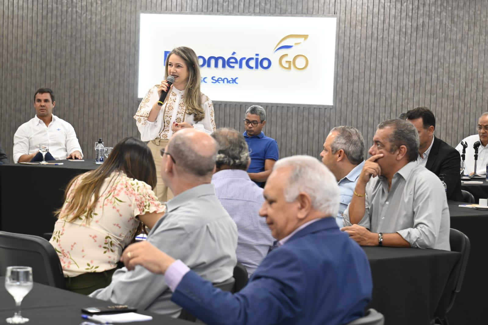 Rejane Duarte em pé, com microfone, conduzindo uma conversa diante de um grupo misto